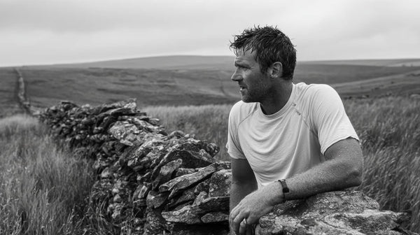  Hiker pausing at drystone wall on Peak District moorland, pulling gently at the back of a plain cotton t-shirt that's slightly damp from a long ascent, overcast British sky