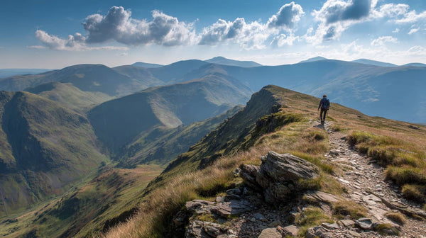 Hiker walking along exposed ridgeline trail in bright summer sunshine, Lake District fells stretching into haze, clear blue sky with scattered cloud, heat shimmer visible on rocky path