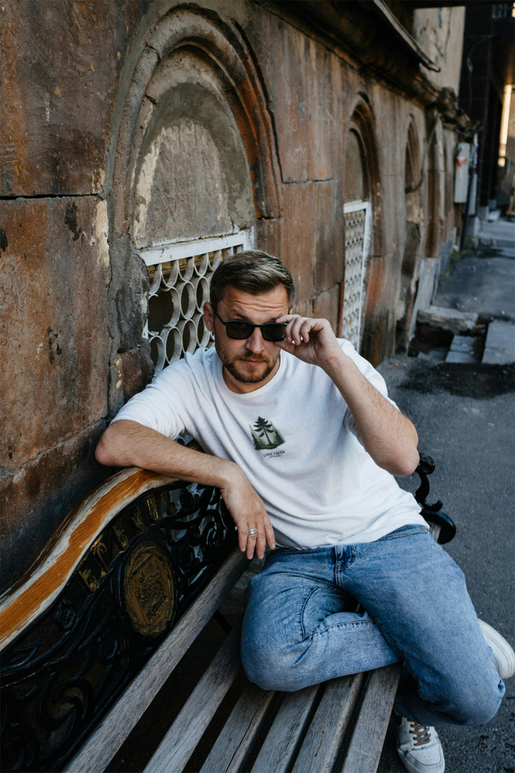 Man sitting on a bench wearing sunglasses and a white t-shirt with a logo.