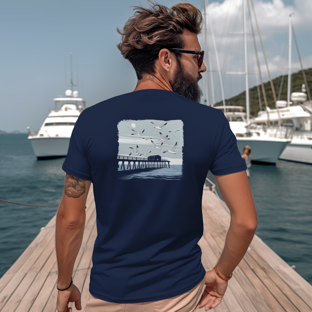 Man wearing a navy blue t-shirt with a graphic design on the back, standing on a dock with boats in the background.
