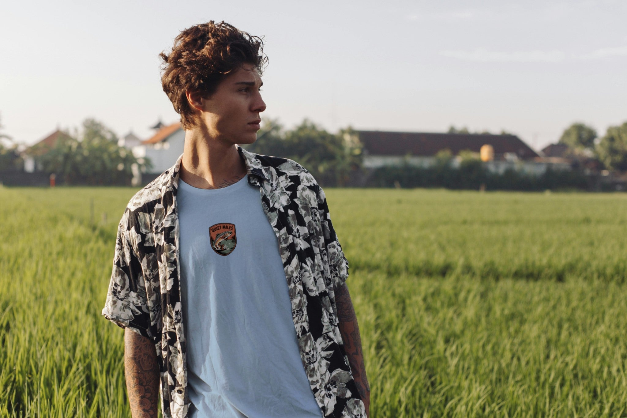 Person standing in a field wearing a patterned shirt over a white t-shirt.