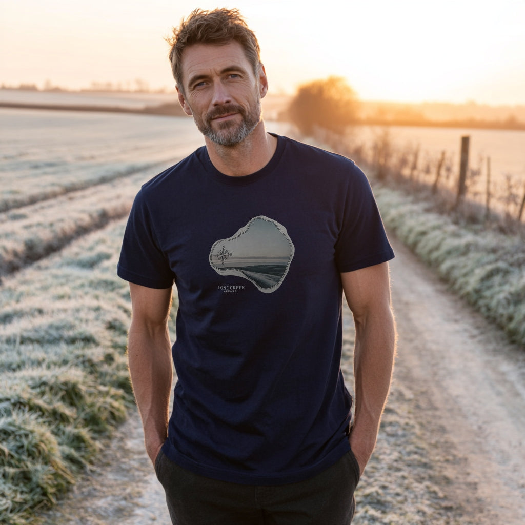 Man wearing a navy blue t-shirt with a graphic design standing in a field at sunset.