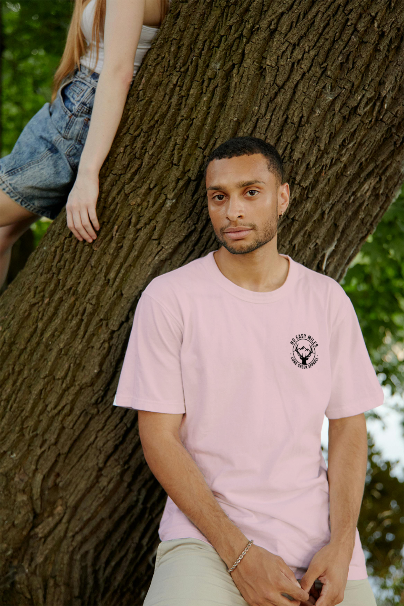 Man wearing a pink t-shirt with a logo, sitting against a tree