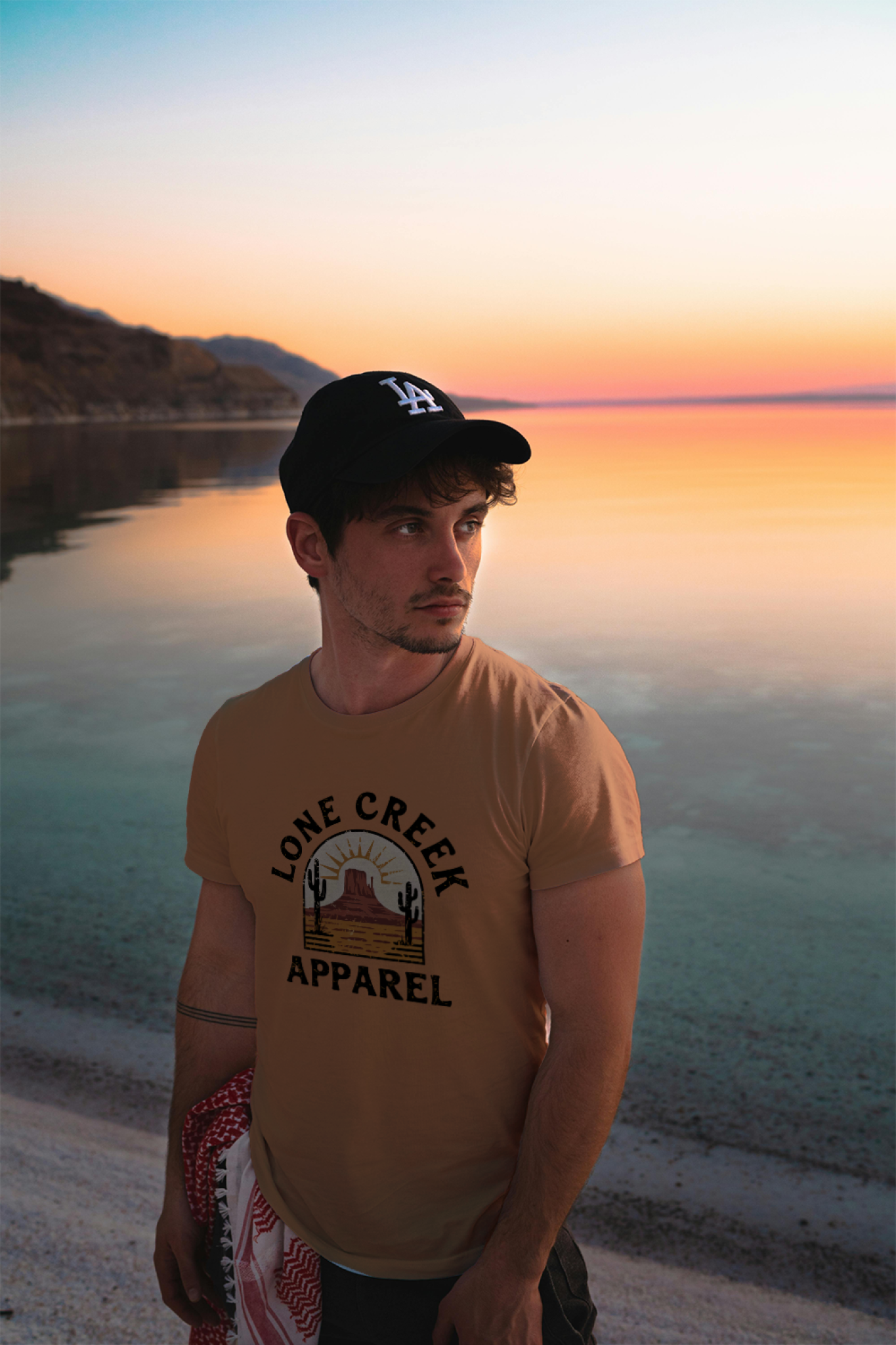 Man wearing a 'Lone Creek Apparel' t-shirt by a lake at sunset